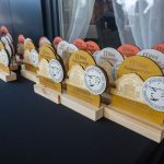 DSC_5350 Na stole stoją drewniane trofea w różnych kolorach z napisami i białymi medalami. Tło jest ciemne z zasłoną. Trofea mają napisy o II Biegu Konstantego.