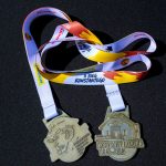 DSC_5353 Dwa medale z białymi wstążkami na czarnym tle. Na jednym medal z twarzą, na drugim budynek i napis Konstanty Łazicki. Kolory wstążek to biały, czerwony i żółty.