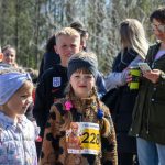 DSC_5360 Grupa dzieci stoi na zewnątrz, niektóre mają numery startowe. Tło to drzewa, a jedna osoba trzyma telefon. Kolory są jasne, wiosenne.