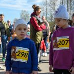 DSC_5362 Na zdjęciu widoczne dzieci w numerach startowych uczestniczą w biegu. Mają na sobie kolorowe bluzy i czapki. Tło stanowi tłum osób na tle drzew.