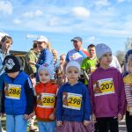 DSC_5365 Dzieci w kolorowych ubraniach z numerami startowymi stoją na zewnątrz. Tło to niebo z chmurami i dorośli w tle. Jest słoneczna pogoda.