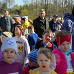 DSC_5367 Grupa dzieci w kolorowych ubraniach stoi na zewnątrz. W tle widać drzewo i rozmyte postacie dorosłych. Dzieci mają numerki na koszulkach, sugerujące udział w biegu.
