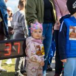 DSC_5370 Na zdjęciu jest małe dziecko w różowej czapce i bluzie z postaciami z bajek. W tle grupa ludzi na zewnątrz. Obok widoczny jest licznik z numerem 37.