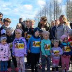DSC_5373 Grupa dzieci w kolorowych ubraniach stoi na zewnątrz z numerami startowymi. Tło stanowią drzewa i niebieskie niebo. W tle dorośli obserwują wydarzenie.