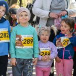 DSC_5376 Grupa dzieci w kolorowych ubraniach z numerami startowymi stoi na zewnątrz. Tło jest rozmyte, co podkreśla głównych bohaterów. Dominują odcienie niebieskiego, różowego i turkusowego.