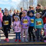 DSC_5388 Grupa dzieci stoi na starcie maratonu, w jasnych kolorowych ubraniach i numerach startowych. Tło to drzewa i niebo. Obraz emanuje energią i radością.