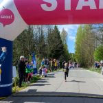 DSC_5393 Start dziecięcego biegu pod różowo-białą bramką z napisem. W tle zielone drzewa i niebieskie niebo. Kibice stoją po bokach.