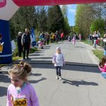 DSC_5405 Grupa dzieci biegnie obok dmuchanej bramy na asfaltowej drodze. Po bokach są widzowie, a zegar pokazuje 10:25. Tło stanowią drzewa i niebo.