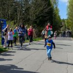 DSC_5410 Dziecko biegnie w wyścigu, otoczone osobami kibicującymi. W tle widać drzewa i niebo. Na przodzie numer startowy 286.