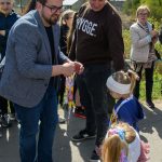 DSC_5412 Na zdjęciu grupa osób na zewnątrz, mężczyzna w okularach wręcza medale dzieciom. Tło to zielone drzewa i jasne niebo. Dzieci mają kolorowe ubrania i wstążki.