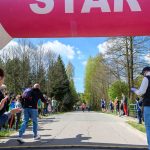 DSC_5435 Na zdjęciu widać start biegowy z wielkim, różowym balonem z napisem START. Zawodnicy biegną po asfaltowej drodze, a widzowie stoją przy trasie wśród drzew. Tło stanowi niebo z białymi chmurami.