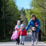 DSC_5455 Na zdjęciu widać trzy osoby spacerujące po ścieżce otoczonej drzewami. Osoba dorosła ubrana w niebieską kurtkę trzyma smartfon. Dwoje dzieci ubranych w kolorowe kurtki idzie obok.