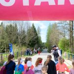 DSC_5465 Na zdjęciu grupa dzieci stoi pod różowym banerem START. W tle widoczna jest droga w otoczeniu drzew. Pogoda jest słoneczna, a przy drodze stoi kilka osób.