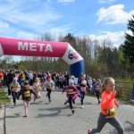 DSC_5483 Dzieci biegną przez różowo-białą bramkę z napisem META. Tło stanowi las i niebieskie niebo z chmurami. Uczestnicy mają na sobie kolorowe stroje i numery startowe.