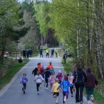 DSC_5488 Dzieci biegają po leśnej drodze, otoczone zielonymi drzewami. Tło stanowi wiosenny las. Ubrania dzieci są kolorowe.