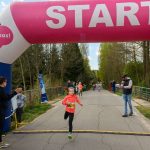 DSC_5497 Dziewczynka w różowej bluzie przekracza linię mety na biegu dziecięcym. Nad nią różowy łuk z napisem START. W tle las i kibice.