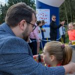 DSC_5500 Mężczyzna w marynarce rozmawia z dziewczynką w stroju sportowym na zawodach. W tle ludzie oraz balon z napisem Ale czas!. Scena odbywa się na zewnątrz, w pobliżu drzew.