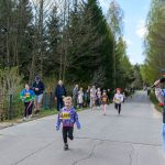 DSC_5507 Dzieci biegają w kolorowych strojach na wiejskiej drodze, otoczonej drzewami. Stojący mężczyzna z mikrofonem prowadzi wydarzenie. Baner reklamowy widoczny z lewej strony drogi.
