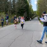DSC_5509 Na zdjęciu widać bieg dzieci na wiejskiej drodze, w tle las. Baner i kibice po lewej, komentator z mikrofonem po prawej. Dzieci mają numery startowe.