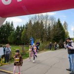DSC_5512 Dzieci biegną pod różowym łukiem z numerami startowymi na jasnej drodze. W tle drzewa i widzowie. Mężczyzna z mikrofonem kibicuje.