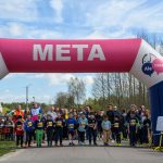 DSC_5523 Ludzie gromadzą się na starcie biegu pod różowo-białym łukiem META. Dzieci w sportowych strojach z numerami na piersi. W tle zielone drzewa i błękitne niebo.