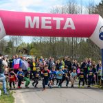 DSC_5525 Rozpoczęcie biegu dzieci na asfaltowej drodze pod różowym balonem z napisem META. Tło to zieleń i drzewa wiosną. Wokół kibice i rodzice.