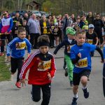 DSC_5537 Młodzi biegacze startują w wyścigu na tle licznej publiczności. Kolorowe stroje dzieci kontrastują z zielenią w tle. Atmosfera sportowego wydarzenia z licznikiem czasowym.