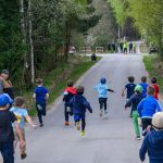 DSC_5542 Dzieci biegną wyścigiem po asfaltowej drodze otoczonej zielonym lasem. Po lewej stronie kilku dorosłych obserwuje wydarzenie. Na końcu drogi widoczni są organizatorzy w kamizelkach.