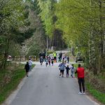 DSC_5546 Droga w lesie z grupą biegnących dzieci i dorosłych. Zielone drzewa i niebo w tle. Ludzie ubrani w kolorowe stroje.