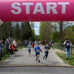 DSC_5551 Dzieci biegną pod różowym łukiem START na leśnej drodze. W tle kibice i drzewa. Na ziemi widoczny jest zegar czasu 0:00:52.