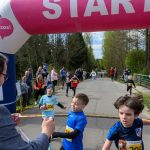 DSC_5556 Dzieci przekraczają linię startową na zawodach biegowych. Tło stanowi zielony las. Nad nimi znajduje się różowy baner „START”.