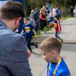 DSC_5558 Na zdjęciu widać grupkę dzieci biegnących alejką. Chłopiec w niebieskiej koszulce z medalem stoi z przodu. W tle ludzie obserwują bieg pośród drzew.