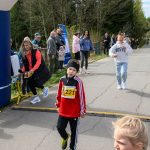 DSC_5559 Uczestnicy biegu na szosie, głównie dzieci, otoczeni drzewami. Żółte numery startowe, ubrania w odcieniach czerwieni i bieli. W tle kibicujący ludzie i niebieski balon startowy.