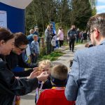 DSC_5565 Grupa osób stoi na ścieżce, dzieci otrzymują medale. W tle zielone drzewa i niebieskie plakaty. Dzień jest słoneczny.