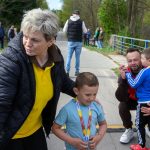 DSC_5567 Na zdjęciu starsza kobieta i dwaj chłopcy z medalami na szyi. Kobieta ma żółtą koszulkę, dzieci są ubrane na niebiesko. W tle widać ludzi i drzewa, chodnik i niebieską poręcz.