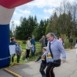 DSC_5568 Kobieta z chłopcem biegnie podczas zawodów, mijając niebieski balon startowy. W tle grupa osób przy drodze. Otoczenie jest zielone z drzewami i trawą.