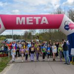 DSC_5573 Grupa dzieci i dorosłych stoi na linii startu biegu pod różowo-niebieskim balonem z napisem META na tle drzew.