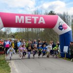 DSC_5576 Dzieci biegają spod różowego nadmuchanego łuku mety. W tle drzewa i widzowie. Niebieska i biała kolorystyka dekoracji.