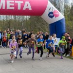 DSC_5580 Grupa dzieci w kolorowych strojach biegnie przez linię mety. Tło stanowi las i tłum kibiców. Nad metą różowy łuk z napisem META.
