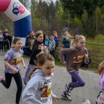 DSC_5584 Grupa dzieci biegnie w wyścigu na tle lasu. Nosi kolorowe koszulki i numery startowe. Za nimi stoi tłum kibiców i duży kolorowy balon.