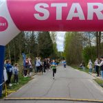 DSC_5589 Biegaczka mija różowy łuk z napisem START. Po bokach stoją drzewa i kibicujący ludzie. Pogoda jest słoneczna.