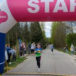 DSC_5590 Biegaczki przebiegają pod różowym łukiem startowym na asfaltowej drodze. Wokół widoczni kibice i zielone drzewa w tle. Pogoda jest pochmurna, atmosfera sportowa.