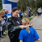 DSC_5595 Na zdjęciu kobieta zakłada medal dziecku w niebieskiej koszulce. W tle ludzie kibicują wydarzeniu na świeżym powietrzu. Dookoła drzewa, asfaltowa droga i plakaty promocyjne.