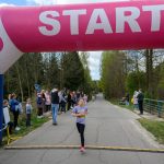 DSC_5599 Na zdjęciu widzimy młodą biegaczkę przekraczającą linię startu. Nad drogą znajduje się różowo-biały łuk z napisem START. W tle widać ludzi obserwujących wydarzenie i zielone drzewa.