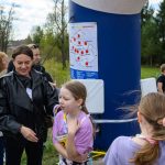 DSC_5603 Grupa osób stoi na zewnątrz przy niebieskim filarze z mapą. Tło to zielone drzewa i wiejskie zabudowania. Dzieci rozmawiają z dorosłymi.