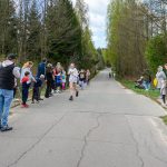 DSC_5604 Na zdjęciu widoczny jest bieg uliczny dzieci i kibice. Przy drodze rosną zielone drzewa. Dzień jest jasny, a niebo lekko zachmurzone.