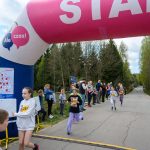 DSC_5606 Na zdjęciu dzieci biegną przez bramę startową podczas wyścigu. Kolory dominujące to różowy i biały na bramie. W tle widoczni są kibice i zalesiona okolica.