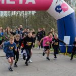 DSC_5619 Dzieci startują w biegu pod różowo-niebieskim łukiem z napisem META. Tło stanowi zielony las. Obok stoi mężczyzna z mikrofonem.