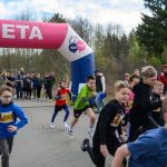 DSC_5622 Młodzież biegnie przez różowy łuk z napisem META. W tle las i grupa kibiców. Dzieci mają różnokolorowe stroje sportowe.
