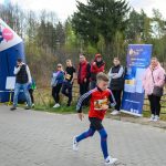 DSC_5625 Chłopiec biegnie w zawodach na tle lasu. Ludzie stoją obok niebiesko-różowego łuku i plakatu. Chłopiec ma czerwoną bluzę i niebieskie legginsy.